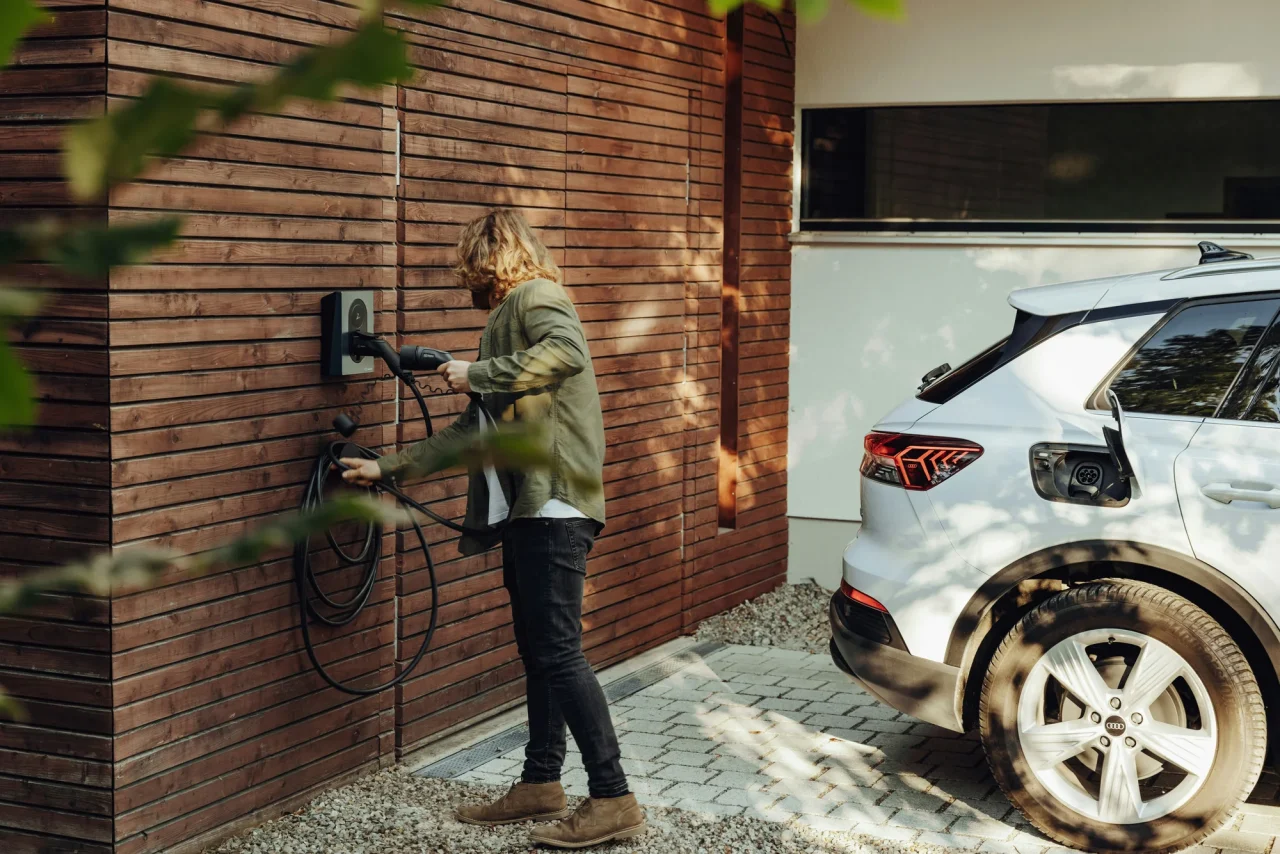 Person charging an electric car outside a timber-clad home.