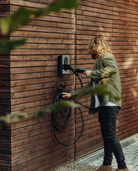 Person charging an electric car outside a timber-clad home.